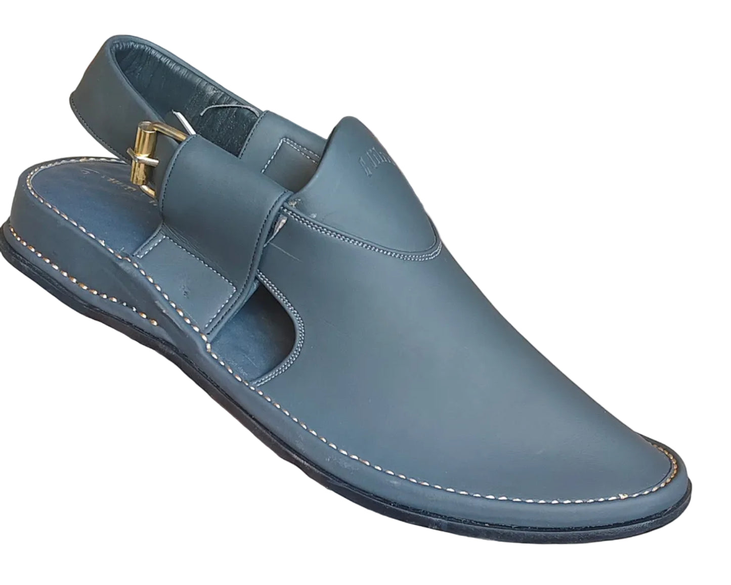 A hand holds a dark slate blue leather men's clog sandal with a gold buckle, set against a rustic brick wall background with other s
hoes visible on the floor.